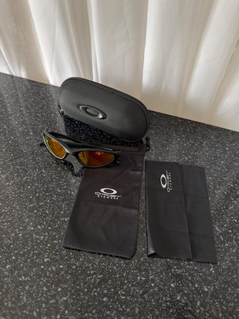 May include: Black sunglasses with orange-tinted lenses, a black carrying case, a black drawstring bag, and a black cleaning cloth. The sunglasses and accessories are from the brand Oakley. The case is oval-shaped.
