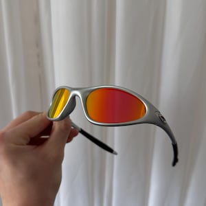 Puede incluir: Gafas de sol plateadas con lentes rojas y amarillas vibrantes. El dise&ntilde;o elegante y moderno presenta una montura curvada y patillas negras. Estas gafas de sol ofrecen un aspecto elegante para actividades al aire libre.