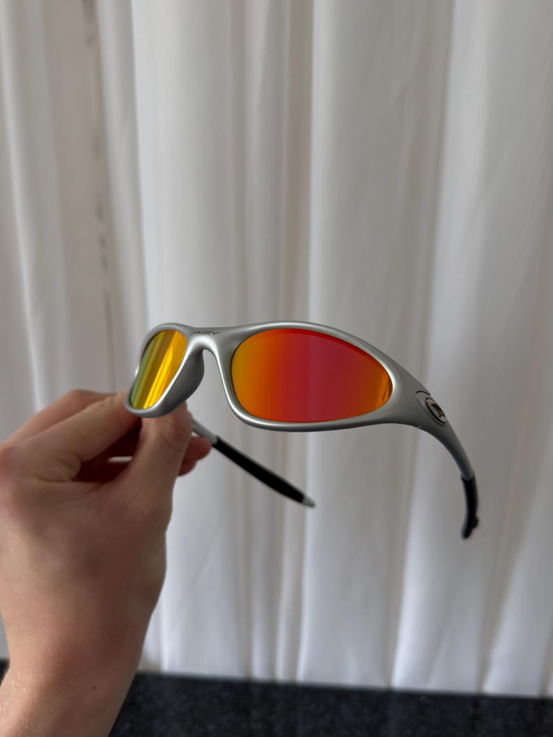 Puede incluir: Gafas de sol plateadas con lentes rojas y amarillas vibrantes. El dise&ntilde;o elegante y moderno presenta una montura curvada y patillas negras. Estas gafas de sol ofrecen un aspecto elegante para actividades al aire libre.