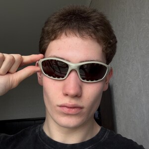 May include: Close-up of silver-framed sunglasses with dark lenses. The sunglasses have a sporty design with a rectangular shape and small ventilation holes. The frame is a light, neutral color, and the lenses are dark, providing contrast.