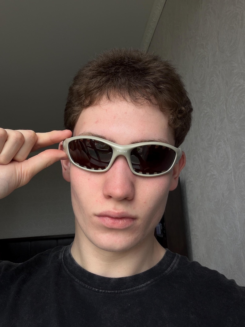 May include: Close-up of silver-framed sunglasses with dark lenses. The sunglasses have a sporty design with a rectangular shape and small ventilation holes. The frame is a light, neutral color, and the lenses are dark, providing contrast.