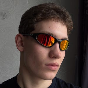 May include: Black sunglasses with a curved frame and orange and red reflective lenses. The sunglasses are worn by a person wearing a black top.