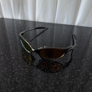 May include: Sleek, rimless sunglasses with a dark gray frame and gradient lenses. The lenses transition from a green hue at the top to a warm amber at the bottom. The sunglasses are resting on a dark, speckled surface.