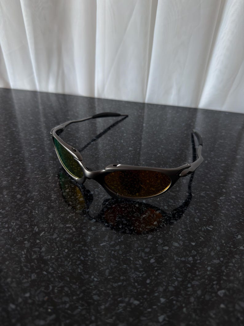 May include: Sleek, rimless sunglasses with a dark gray frame and gradient lenses. The lenses transition from a green hue at the top to a warm amber at the bottom. The sunglasses are resting on a dark, speckled surface.