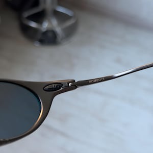May include: Close-up of a pair of sunglasses with a dark gray frame and blue lenses. The text "ROMEO CE" is printed on the temple arm. The sunglasses are set against a blurred background.
