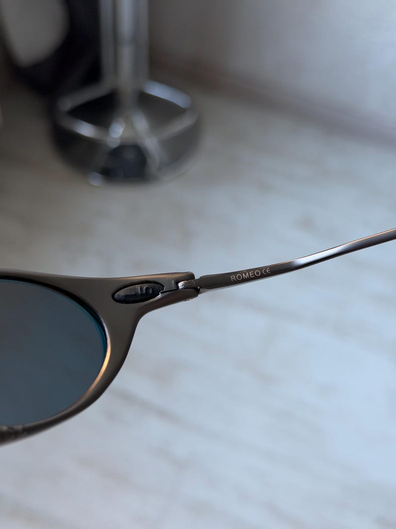 May include: Close-up of a pair of sunglasses with a dark gray frame and blue lenses. The text "ROMEO CE" is printed on the temple arm. The sunglasses are set against a blurred background.