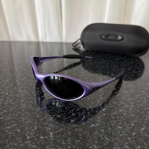 May include: Purple sunglasses with black lenses and a matching black case. The sunglasses have a sleek, curved design. The case is oval-shaped with a zipper closure and a small logo.