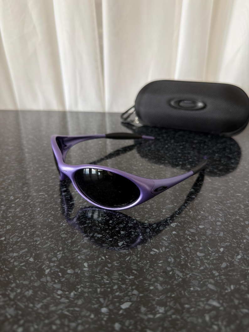 May include: Purple sunglasses with black lenses and a matching black case. The sunglasses have a sleek, curved design. The case is oval-shaped with a zipper closure and a small logo.