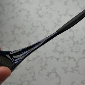 K&ouml;nnte beinhalten: Nahaufnahme einer schwarzen Sonnenbrille mit blau schillerndem Finish. Der B&uuml;gel ist sichtbar, mit dem Text "MADE IN U.S.A." in den Rahmen ge&auml;tzt. Die Sonnenbrille hat ein schlankes, modernes Design.
