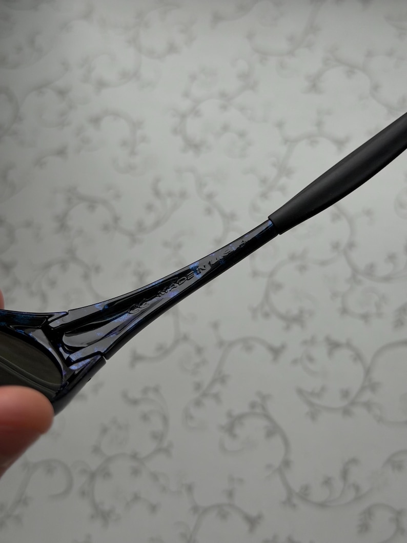 K&ouml;nnte beinhalten: Nahaufnahme einer schwarzen Sonnenbrille mit blau schillerndem Finish. Der B&uuml;gel ist sichtbar, mit dem Text "MADE IN U.S.A." in den Rahmen ge&auml;tzt. Die Sonnenbrille hat ein schlankes, modernes Design.