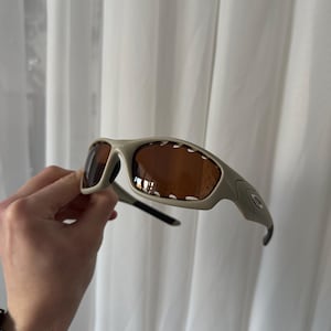 May include: Tan-colored sunglasses with brown lenses. The sunglasses have a curved frame and a sporty design. The Oakley logo is visible on the side of the frame. The sunglasses are held against a white curtain background.