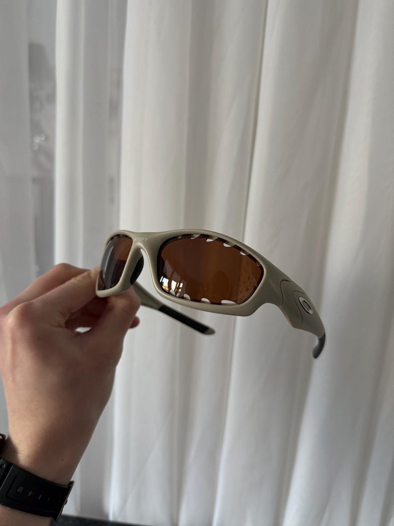 May include: Tan-colored sunglasses with brown lenses. The sunglasses have a curved frame and a sporty design. The Oakley logo is visible on the side of the frame. The sunglasses are held against a white curtain background.