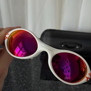 May include: White sunglasses with a distinctive oval frame and vibrant red-purple lenses. The Oakley logo is visible on the frame. The temples have a red accent. A black case is in the background.