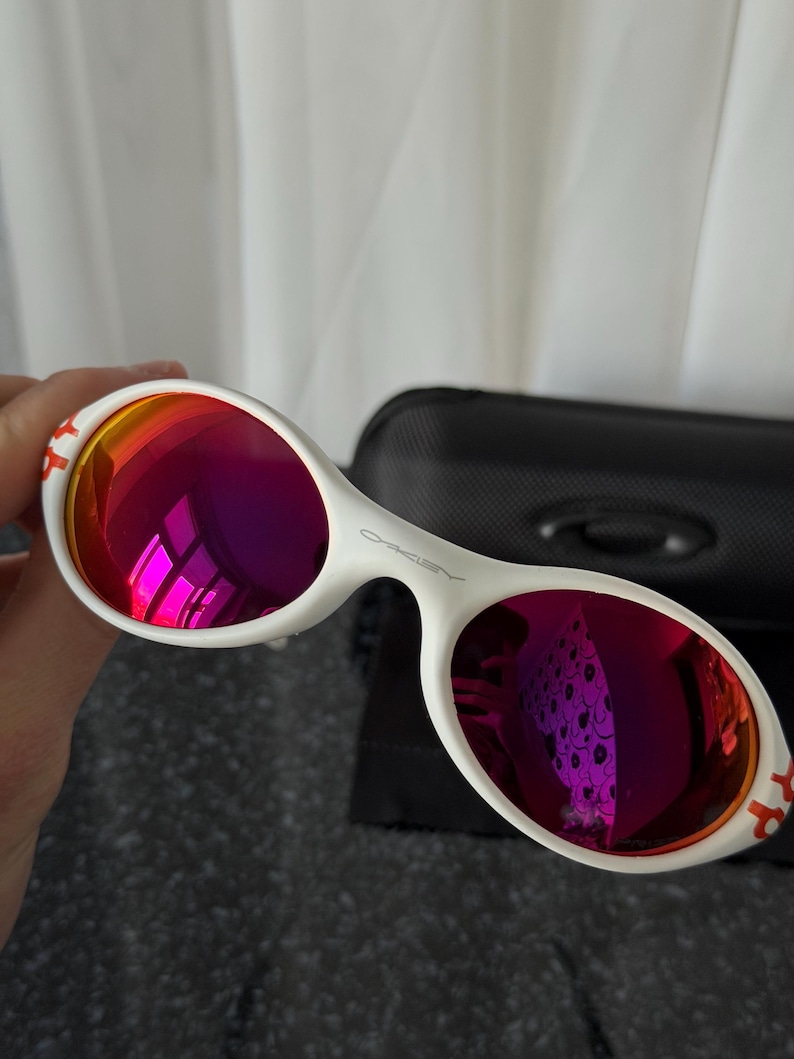 May include: White sunglasses with a distinctive oval frame and vibrant red-purple lenses. The Oakley logo is visible on the frame. The temples have a red accent. A black case is in the background.