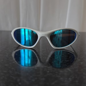 Puede incluir: Gafas de sol con montura blanca y lentes azules reflectantes. Las gafas de sol est&aacute;n sobre una superficie oscura y reflectante, con una cortina blanca suave en el fondo. Las lentes reflejan un tono azul brillante.