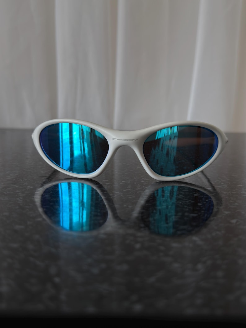 Puede incluir: Gafas de sol con montura blanca y lentes azules reflectantes. Las gafas de sol est&aacute;n sobre una superficie oscura y reflectante, con una cortina blanca suave en el fondo. Las lentes reflejan un tono azul brillante.