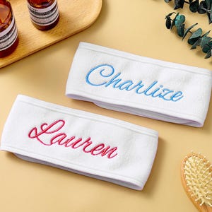 May include: Two white terry cloth headbands embroidered with names in cursive. One headband has "Charlize" in blue, the other has "Lauren" in pink. The headbands are on a light yellow surface, with brown bottles and a wooden tray in the background.