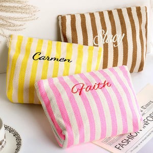 May include: Three striped pouches in yellow, brown, and pink, each with a different name embroidered on them: "Carmen", "Stacy", and "Faith". The pouches are rectangular and have a zipper closure. The background includes a cup and saucer, and a newspaper.
