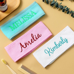 Op de afbeelding: Drie gepersonaliseerde spa-hoofdbanden in teal, roze en wit, elk geborduurd met een andere naam in een contrasterende kleur. De hoofdbanden zijn gemaakt van een textuurstof op een lichtgele ondergrond.