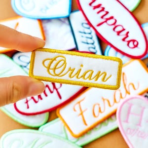 Könnte beinhalten: Ein rechteckiger weißer Aufnäher mit dem Namen "Orian" in goldener Schrift, gehalten von einer Hand. Der Aufnäher hat einen goldenen Rand. Im Hintergrund befinden sich weitere bestickte Namensaufnäher in verschiedenen Formen und Farben.