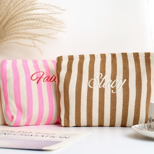May include: Two striped cosmetic bags. One is pink and white with "Fair" embroidered in red. The other is brown and white with "Stacy" embroidered in white. The bags are rectangular and sit on a white surface.