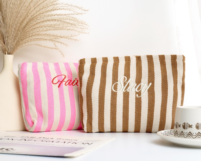 May include: Two striped cosmetic bags. One is pink and white with "Fair" embroidered in red. The other is brown and white with "Stacy" embroidered in white. The bags are rectangular and sit on a white surface.