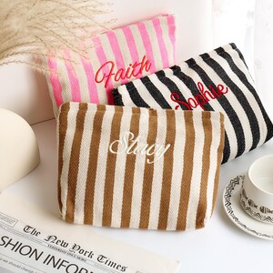 May include: Three striped canvas pouches in pink, black and white, and brown and white. Each pouch has a name embroidered on it in a contrasting color: "Faith" in red, "Sophie" in red, and "Stacy" in white. A cup and saucer are in the background.