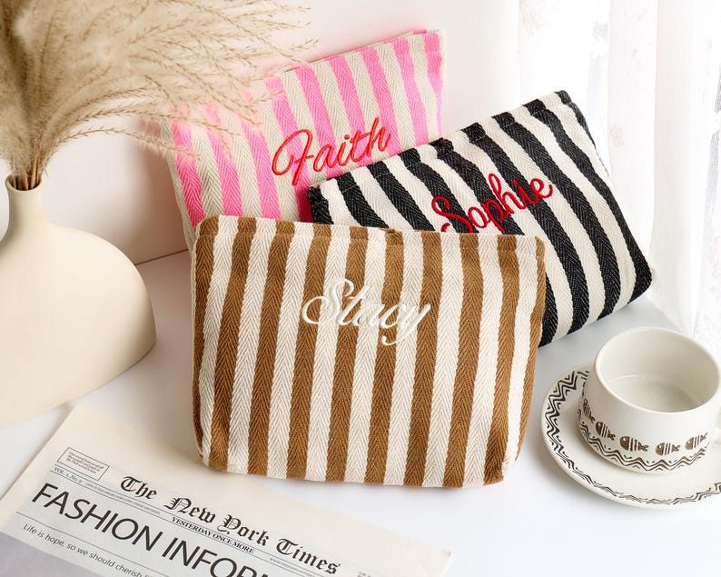 May include: Three striped canvas pouches in pink, black and white, and brown and white. Each pouch has a name embroidered on it in a contrasting color: "Faith" in red, "Sophie" in red, and "Stacy" in white. A cup and saucer are in the background.