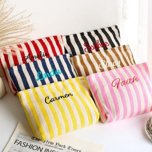 May include: A collection of personalized striped pouches in various colors, including red, black, blue, brown, yellow, and pink. Each pouch features a different name embroidered in a contrasting color. The pouches are arranged on a white surface.
