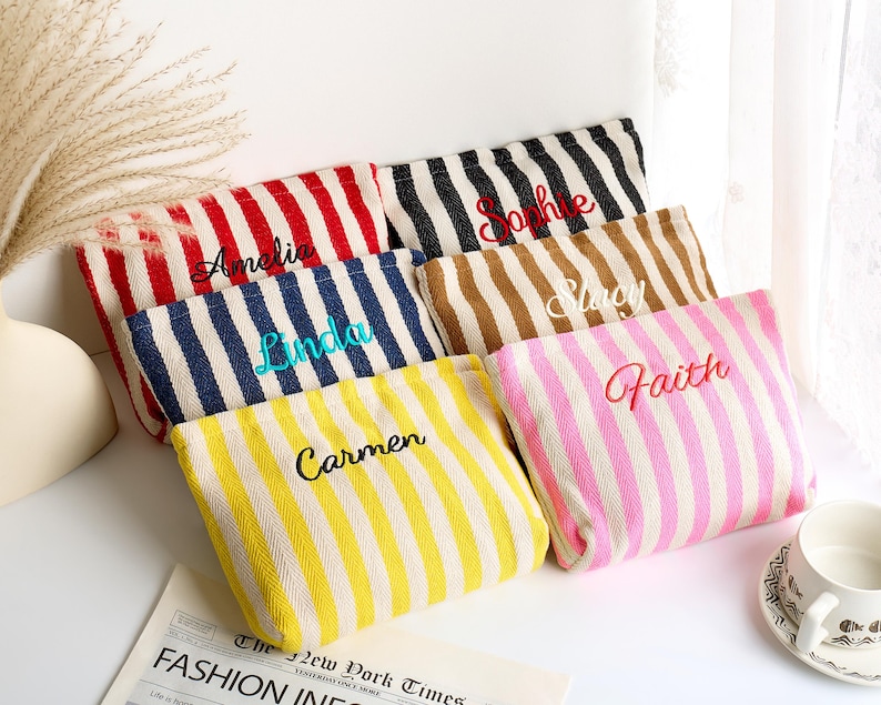 May include: A collection of personalized striped pouches in various colors, including red, black, blue, brown, yellow, and pink. Each pouch features a different name embroidered in a contrasting color. The pouches are arranged on a white surface.