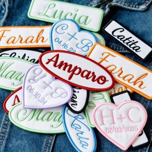 Puede incluir: Una colección de parches bordados con nombres en varias formas y colores, incluyendo rectángulos y corazones. Los parches presentan nombres como "Lucia", "Farah" y "Amparo", junto con fechas e iniciales, todos cosidos en colores de hilo contrastantes sobre un fondo blanco.