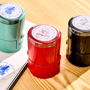 Op de afbeelding: Drie zelfinktende bibliotheekstempels in teal, rood en zwart. Elke stempel heeft een cirkelvormig ontwerp met de tekst "FROM THE LIBRARY OF" en een naam. Een afdruk is zichtbaar op een notitieboekpagina. De stempels staan op een houten oppervlak.