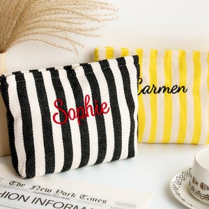 May include: Two striped cosmetic bags. One is black and white with "Sophie" embroidered in red. The other is yellow and white with "Carmen" embroidered in black. The bags are on a white surface with a newspaper and a cup.