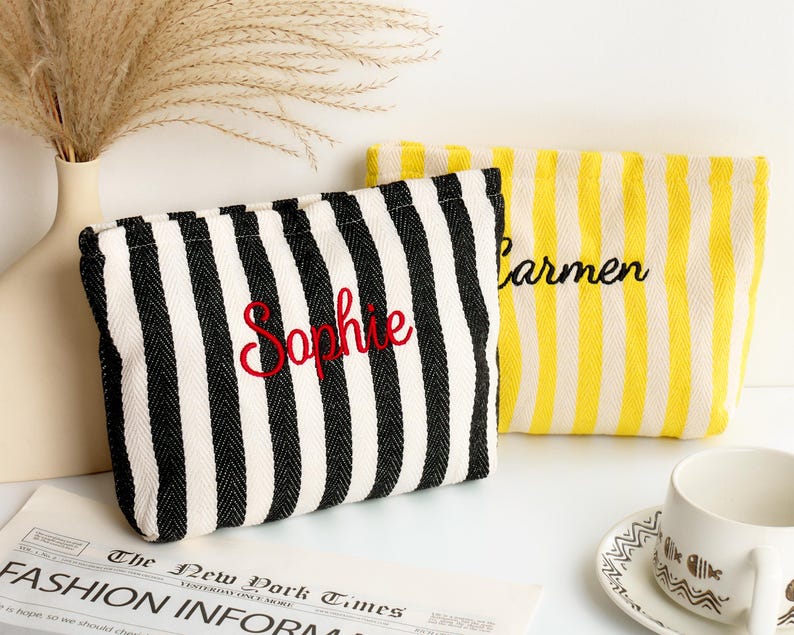 May include: Two striped cosmetic bags. One is black and white with "Sophie" embroidered in red. The other is yellow and white with "Carmen" embroidered in black. The bags are on a white surface with a newspaper and a cup.