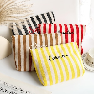 May include: Four striped canvas pouches in black and white, red and white, brown and white, and yellow and white. Each pouch has a name embroidered in black thread: Sophie, Amelia, Stacy, and Carmen. The pouches are arranged on a white surface.