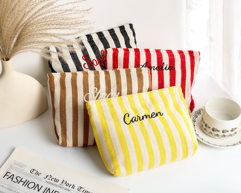 May include: Four striped canvas pouches in black and white, red and white, brown and white, and yellow and white. Each pouch has a name embroidered in black thread: Sophie, Amelia, Stacy, and Carmen. The pouches are arranged on a white surface.