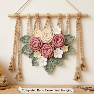 Op de afbeelding: Een wandhanger bestaande uit een houten stok met hangende beige gevlochten koorden en kwastjes. Aan de koorden is een gehaakt bloemstuk bevestigd met oudroze en crèmekleurige rozen, witte bloemen en gehaakte bladeren in saliegroen.