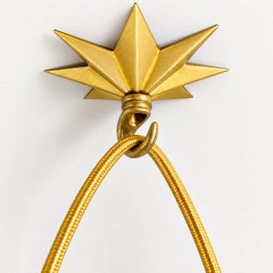 May include: A gold-colored metal wall hook featuring a starburst or sunburst design with multiple pointed rays. A curved hook extends from the center of the starburst, supporting a textured, gold-colored fabric-covered electrical cord.