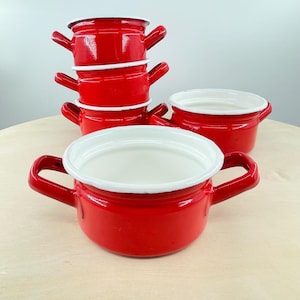May include: Five small, red enamel pots with white rims and handles are stacked. Two more pots are visible. The pots are on a light-coloured wooden surface. The pots are suitable for cooking.