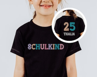 Op de afbeelding: Zwart T-shirt met het woord "SCHULKIND" in kleurrijke, getextureerde letters op de voorkant. De achterkant van het shirt heeft het nummer "25" en de naam "THALIA".
