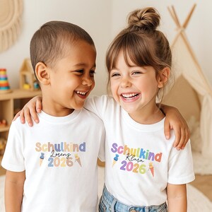 May include: Two children wearing white t-shirts with the text "Schulkind" and a name, plus the year 2026 in colorful letters. The children are smiling and embracing each other. The background includes a wooden shelf with toys and a small teepee.