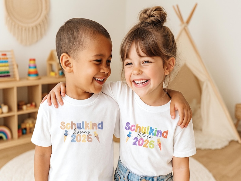 May include: Two children wearing white t-shirts with the text "Schulkind" and a name, plus the year 2026 in colorful letters. The children are smiling and embracing each other. The background includes a wooden shelf with toys and a small teepee.