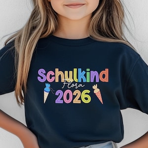 May include: A navy blue t-shirt with the words "Schukind Flora 2026" in colorful letters. The design includes ice cream cones and a carrot. The shirt is likely for a child.