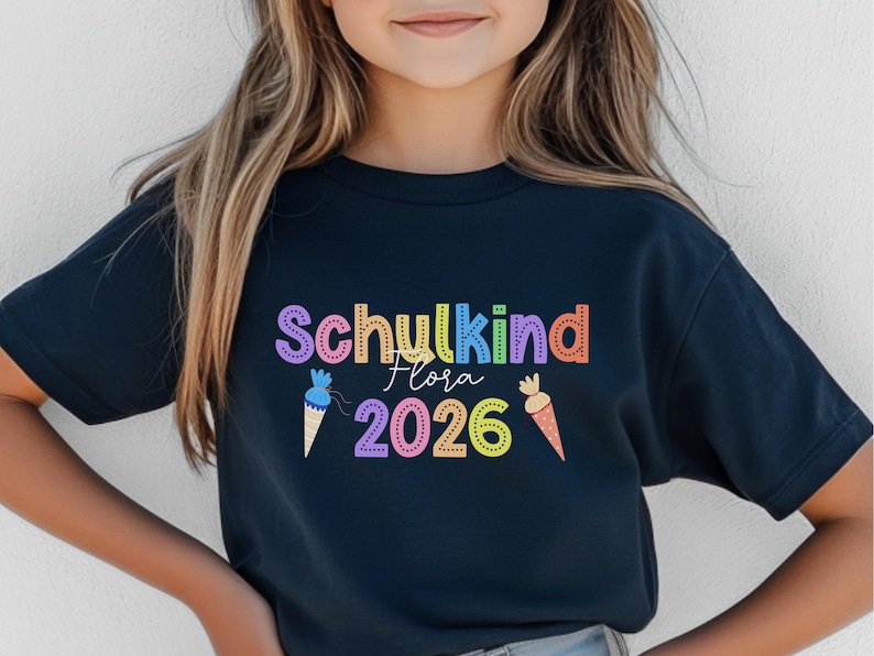 May include: A navy blue t-shirt with the words "Schukind Flora 2026" in colorful letters. The design includes ice cream cones and a carrot. The shirt is likely for a child.