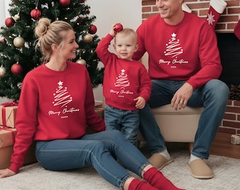 May include: Red Christmas sweatshirts with a white Christmas tree design and the words "Merry Christmas." The sweatshirts are worn by a family of three. The mother is sitting on the floor, the father is sitting in a chair, and the child is standing. The sweatshirts have the words "Mama," "Papa," and "Liam" on them.