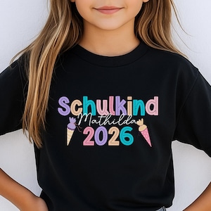 May include: Black t-shirt with the text "Schulkind Matilda 2026" in colorful bubble letters and ice cream cone graphics. The shirt is a crew neck and appears to be made of cotton or a similar fabric.