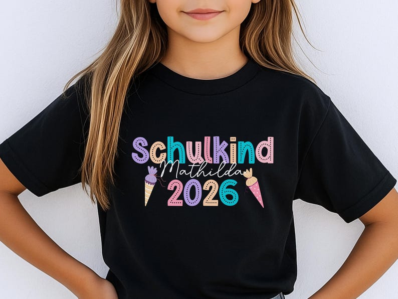 May include: Black t-shirt with the text "Schulkind Matilda 2026" in colorful bubble letters and ice cream cone graphics. The shirt is a crew neck and appears to be made of cotton or a similar fabric.