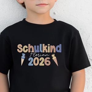 May include: Black t-shirt with the text "Schulkind Florian 2026" in colorful letters. The design includes ice cream cone graphics. The shirt is made of a soft material and is suitable for children.