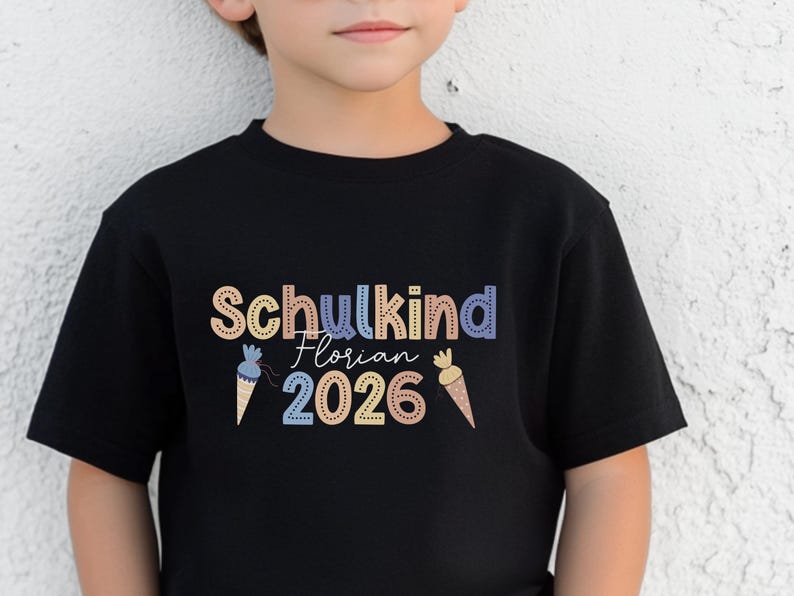 May include: Black t-shirt with the text "Schulkind Florian 2026" in colorful letters. The design includes ice cream cone graphics. The shirt is made of a soft material and is suitable for children.