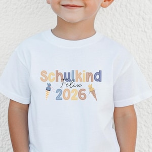 May include: White t-shirt with the text "Schulkind Felix 2026" in colorful letters and decorative ice cream cone graphics. The shirt is made of a soft material and is suitable for children.